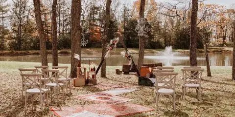 Outdoor wedding ceremony setup with a floral arch, rustic chairs, and rugs on a lawn near a pond.