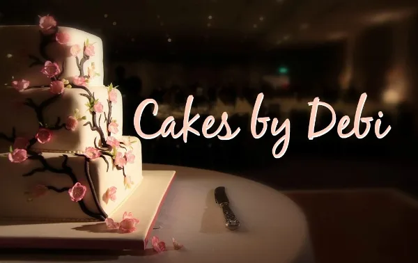 A three-tiered white wedding cake decorated with pink flowers and dark branches, sitting on a table with a cake knife.