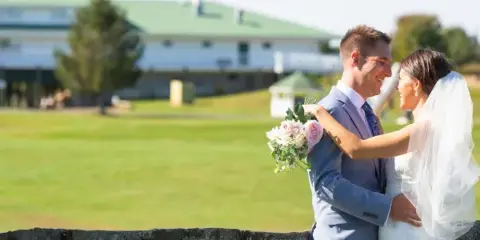 A bride and groom standing outdoors on a golf course with a large white building in the background.