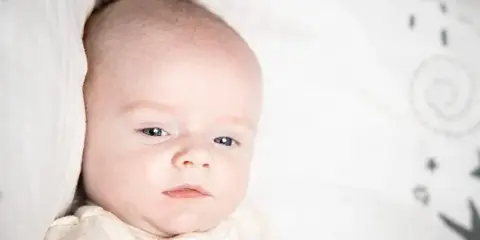 A professional close-up portrait of a newborn baby lying on a white textured blanket with a soft-focus background.