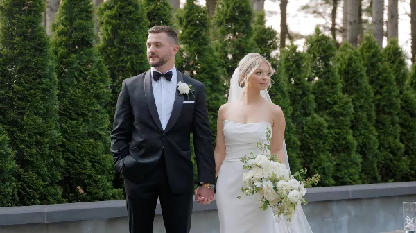 A bride and groom standing together outdoors in formal wedding attire, holding hands in front of a green hedge.
