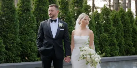 A bride and groom standing together outdoors in formal wedding attire, holding hands in front of a green hedge.