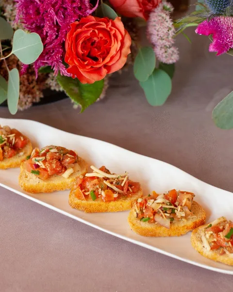 A white platter of bruschetta topped with tomatoes and cheese, set against a backdrop of a vibrant floral arrangement.