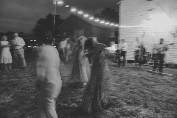 Black-and-white photo of a couple dancing at an outdoor wedding reception with guests and a live band under string lights.