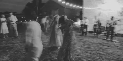 Black-and-white photo of a couple dancing at an outdoor wedding reception with guests and a live band under string lights.
