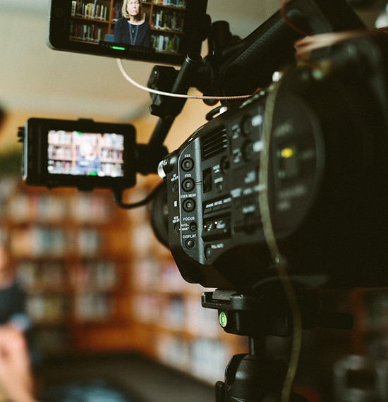 A professional video camera with external monitors is set up to film a person in a room with bookshelves.
