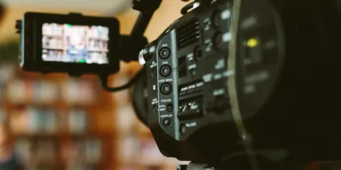 A professional video camera with external monitors is set up to film a person in a room with bookshelves.