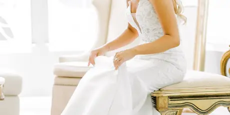 A bride in a white wedding dress sitting on a gold-trimmed bench in a bright room.