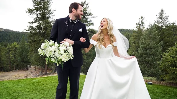 A bride and groom standing together outdoors in a grassy area with trees in the background.