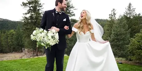 A bride and groom standing together outdoors in a grassy area with trees in the background.