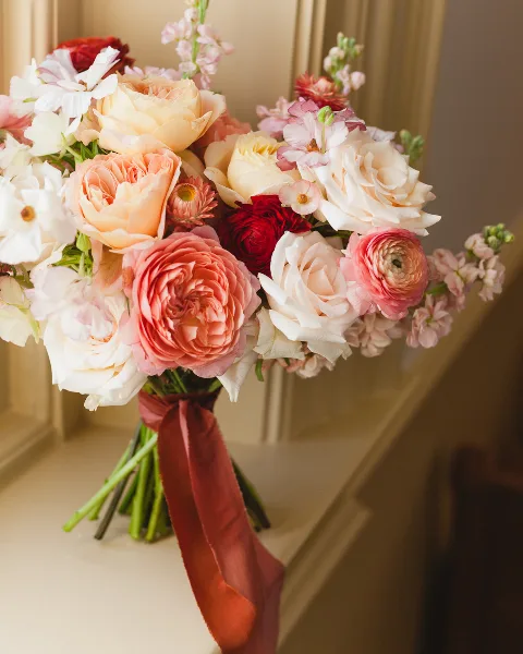A close-up of a romantic bridal bouquet featuring pink and white roses, ranunculus, and other flowers tied with a red ribbon.