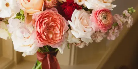 A close-up of a romantic bridal bouquet featuring pink and white roses, ranunculus, and other flowers tied with a red ribbon.