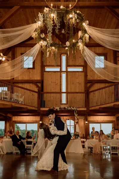 A bride and groom dancing at a wedding reception inside a rustic wooden barn venue with a floral chandelier and draped fabric.