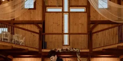 A bride and groom dancing at a wedding reception inside a rustic wooden barn venue with a floral chandelier and draped fabric.