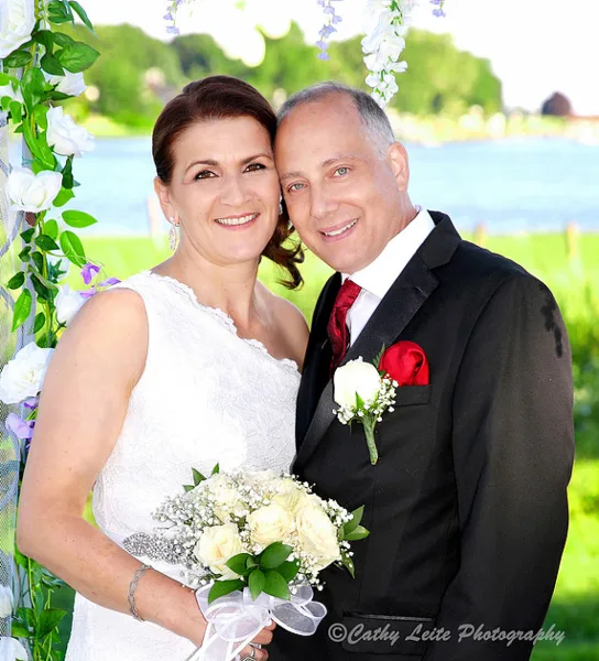 A bride and groom posing together outdoors with a white rose bouquet and floral arch.