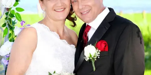 A bride and groom posing together outdoors with a white rose bouquet and floral arch.