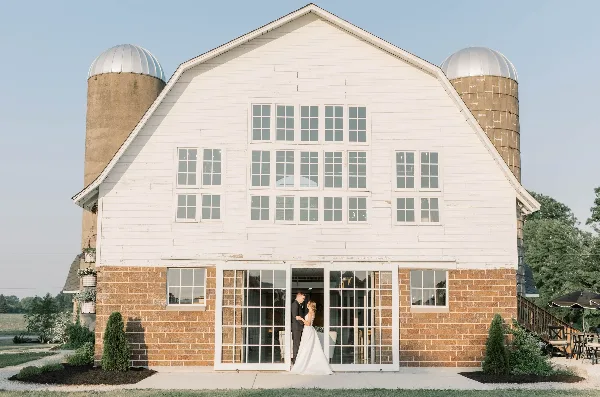 Exterior view of a white barn wedding venue with silos and a bride and groom standing at the entrance.