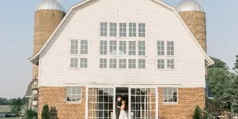 Exterior view of a white barn wedding venue with silos and a bride and groom standing at the entrance.
