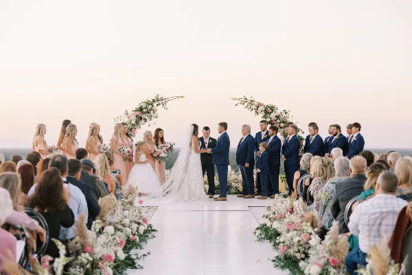 A wide shot of a wedding ceremony taking place outdoors at sunset, featuring a bride and groom exchanging vows under a floral arch with their wedding party and guests.