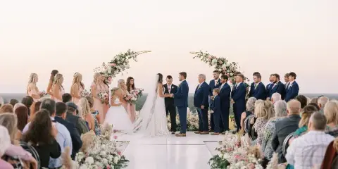 A wide shot of a wedding ceremony taking place outdoors at sunset, featuring a bride and groom exchanging vows under a floral arch with their wedding party and guests.
