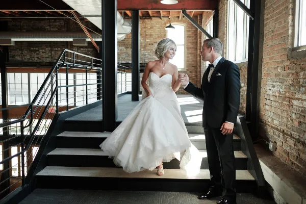 A bride in a white gown and a groom in a tuxedo standing on a staircase in a brick-walled venue.