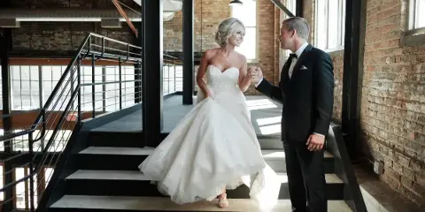 A bride in a white gown and a groom in a tuxedo standing on a staircase in a brick-walled venue.