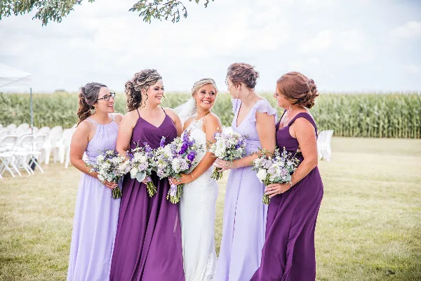 A bride and four bridesmaids in purple and lavender dresses holding bouquets, standing outdoors in a field with white chairs and a cornfield in the background.