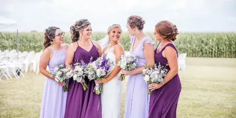 A bride and four bridesmaids in purple and lavender dresses holding bouquets, standing outdoors in a field with white chairs and a cornfield in the background.