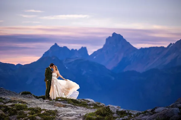 A bride and groom kissing on a rocky mountain summit with a dramatic mountain range and sunset sky in the background.