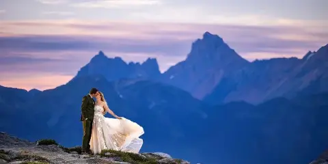 A bride and groom kissing on a rocky mountain summit with a dramatic mountain range and sunset sky in the background.