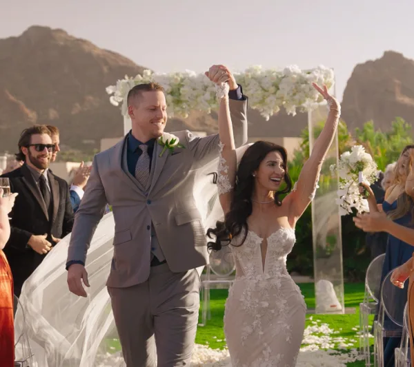 A bride and groom walking down the aisle with arms raised in celebration at an outdoor wedding venue.