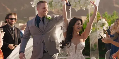 A bride and groom walking down the aisle with arms raised in celebration at an outdoor wedding venue.