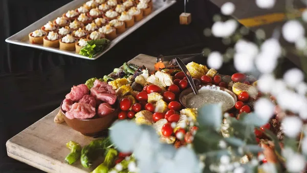 A catering table featuring a large wooden board with a charcuterie spread of meats, roasted vegetables, and a white platter of individual appetizers.