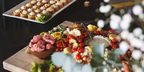 A catering table featuring a large wooden board with a charcuterie spread of meats, roasted vegetables, and a white platter of individual appetizers.