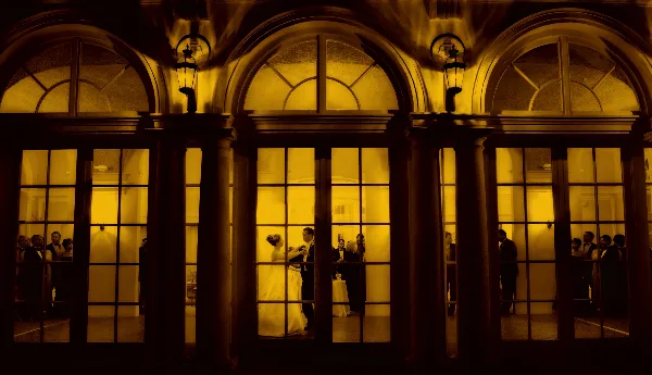 A bride and groom dancing in a grand, warmly lit ballroom with arched windows and guests in the background.