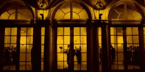 A bride and groom dancing in a grand, warmly lit ballroom with arched windows and guests in the background.