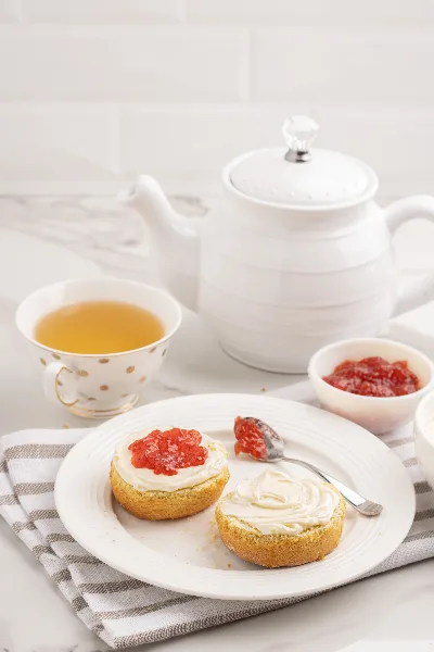 A plate of split scones topped with cream and jam, served with a cup of tea and a white ceramic teapot.