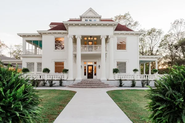 Exterior view of a large white historic mansion with a red roof, columns, and a paved walkway leading to the entrance.