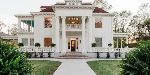 Exterior view of a large white historic mansion with a red roof, columns, and a paved walkway leading to the entrance.