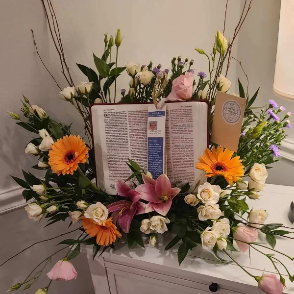 A floral arrangement with orange gerberas, pink lilies, and white roses surrounding an open book on a white surface.