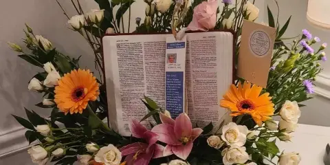 A floral arrangement with orange gerberas, pink lilies, and white roses surrounding an open book on a white surface.