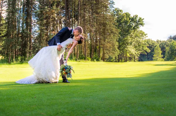 A bride and groom kissing on a green golf course with a forest background.