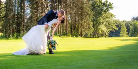 A bride and groom kissing on a green golf course with a forest background.