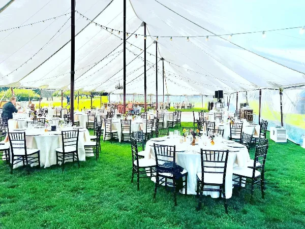 A large white event tent set up on a lawn with multiple round tables dressed in white linens, surrounded by black Chiavari chairs and string lights.