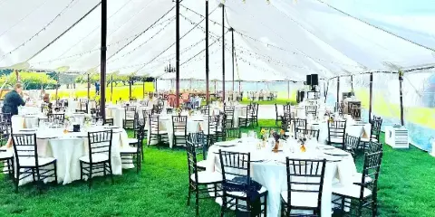 A large white event tent set up on a lawn with multiple round tables dressed in white linens, surrounded by black Chiavari chairs and string lights.