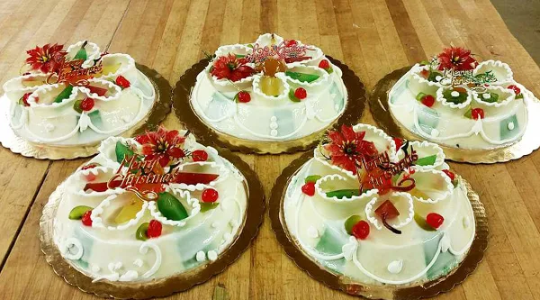 Five decorated Italian specialty cakes with white icing, candied fruit, and red flowers arranged on a wooden table.