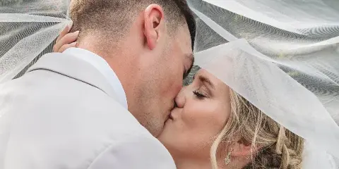A bride and groom kissing under a white wedding veil.