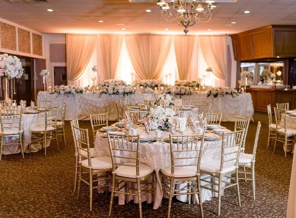 A wedding reception room featuring gold chiavari chairs, tables with lace linens and floral centerpieces, and a backdrop with peach drapery.