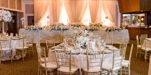 A wedding reception room featuring gold chiavari chairs, tables with lace linens and floral centerpieces, and a backdrop with peach drapery.