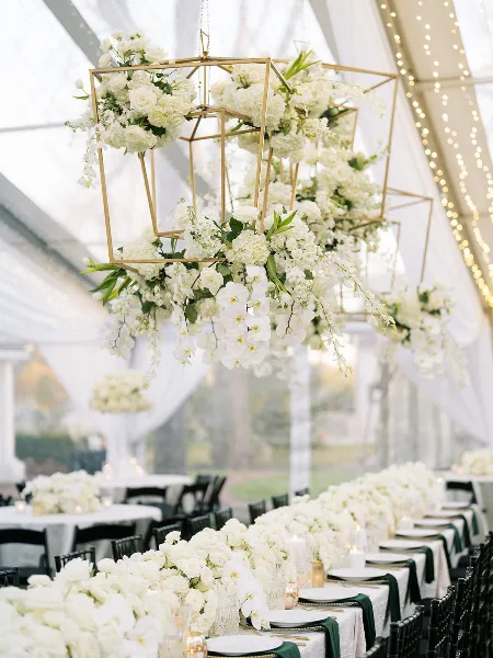 Hanging geometric floral chandeliers and long white table centerpieces in a wedding tent.